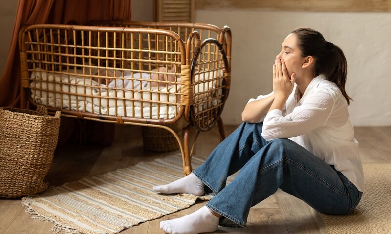 Erschöpfte Mutter mit Wochenbettdepression sitzt neben dem Kinderbett ihres Babys